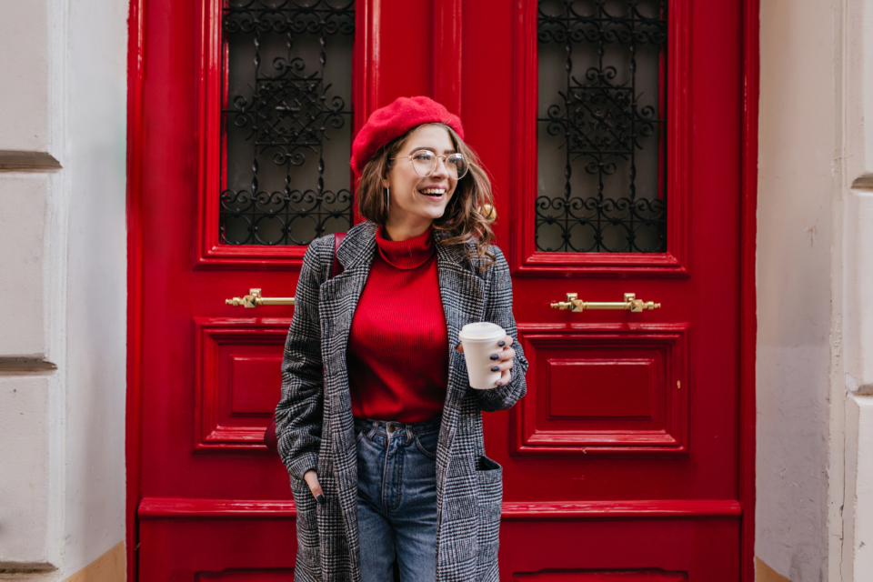 Glad white female model in long gray jacket looking away with smile standing near red door. Outdoor photo of beautiful caucasian woman in sweater drinks coffee in front of cafe and laughing.