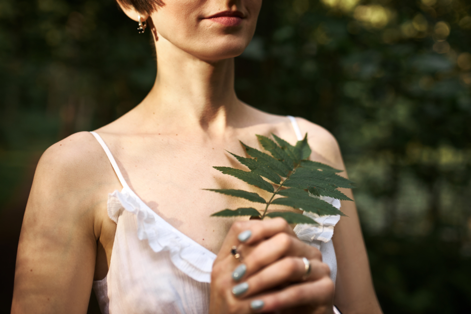 Unrecognizable mysterious young woman with short hairstyle and pale skin walking in forest alone, holding green fern leaf. People, flora, botany and nature concept. Selective focus on female hands